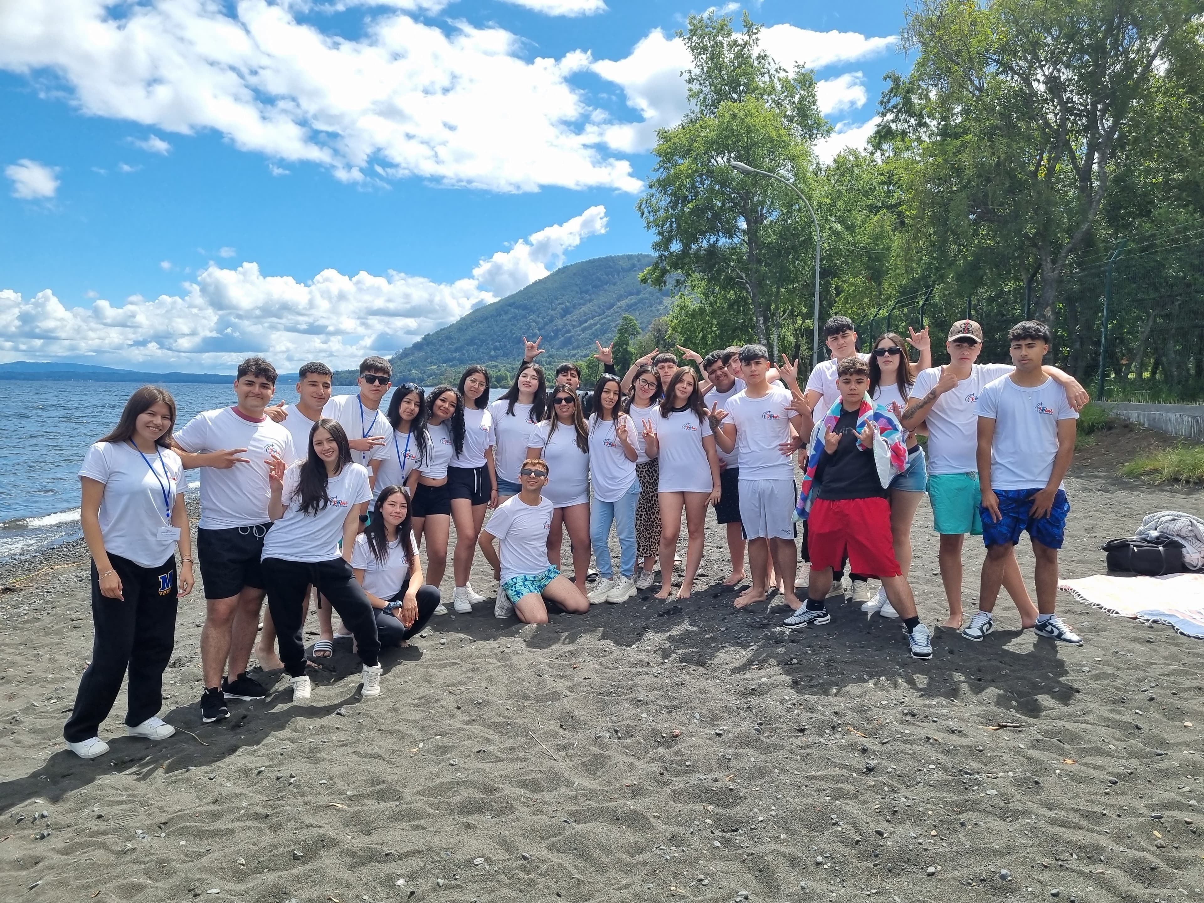 Grupo de estudiantes en gira de estudio posando en playa de arena volcánica con lago y montañas al fondo
